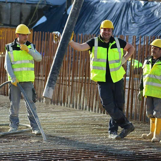 Drei Bauarbeiter in Warnwesten und Helmen arbeiten auf einer Baustelle mit Betonstahl und einer Betonschlauchleitung.